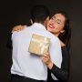 Thankful beautiful brunette young woman in nice elegant dress with christmas gift box hugging her husband or boyfriend and smiling, loving couple celebrating xmas on black background